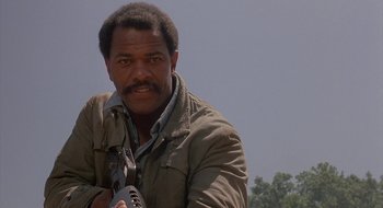 Movie still from “Commando” (1985), directed by Mark L. Lester – A man holding a gun while standing in a field; Close Up shot, Low angle