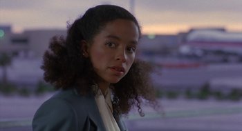 Movie still from “Commando” (1985), directed by Mark L. Lester – A young woman with curly black hair standing in a parking lot at dusk; Close Up shot, Over the shoulder angle