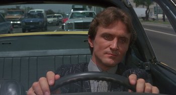 Movie still from “Commando” (1985), directed by Mark L. Lester – A man sitting in the drivers seat of a car while holding a steering wheel; Close Up shot, Low angle