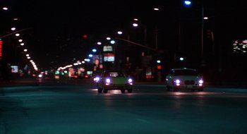Movie still from “Commando” (1985), directed by Mark L. Lester – Cars driving down a street at night with lights on; Wide shot, Low angle