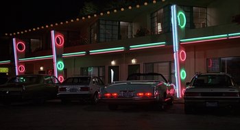 Movie still from “Commando” (1985), directed by Mark L. Lester – Two cars are parked in front of a building; Wide shot, Low angle