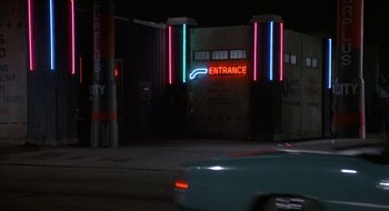 Movie still from “Commando” (1985), directed by Mark L. Lester – A car driving down a street next to a building; Wide shot, Low angle