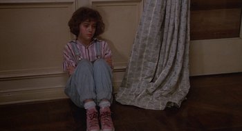 Movie still from “Commando” (1985), directed by Mark L. Lester – A girl sitting on the floor in front of a curtain; Medium shot, High angle