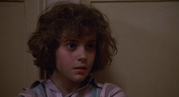 Movie still from “Commando” (1985), directed by Mark L. Lester – A young girl with brown curly hair sitting in front of a wall; Close Up shot, Low angle