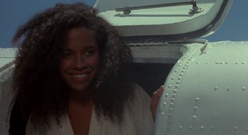 Movie still from “Commando” (1985), directed by Mark L. Lester – A woman with curly black hair smiling for the camera; Close Up shot, Over the shoulder angle