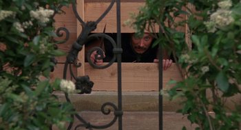 Movie still from “Commando” (1985), directed by Mark L. Lester – A man looking out a window through a metal fence; Wide shot, High angle