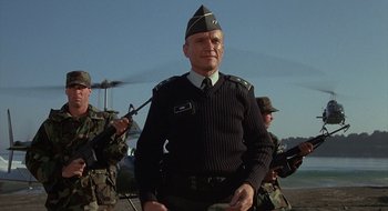 Movie still from “Commando” (1985), directed by Mark L. Lester – An older man in a uniform is posing for a picture; Medium shot, Low angle