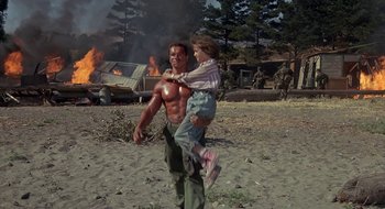 Movie still from “Commando” (1985), directed by Mark L. Lester – A man carrying a young girl on his back in front of a burning building; Wide shot, Low angle