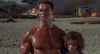 Movie still from “Commando” (1985), directed by Mark L. Lester – A man and a boy are standing in front of a burning building; Medium shot, Low angle