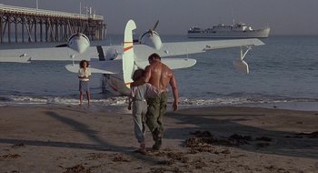 Movie still from “Commando” (1985), directed by Mark L. Lester – A man walking on the beach next to a woman; Wide shot, Low angle