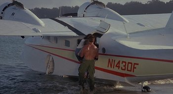 Movie still from “Commando” (1985), directed by Mark L. Lester – A man standing in front of an airplane in the water; Wide shot, Low angle