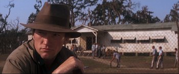 Movie still from “Cool Hand Luke” (1967), directed by Stuart Rosenberg – A man in a hat is holding a baseball bat in front of a house; Close Up shot, Over the shoulder angle