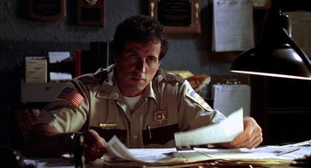 Movie still from “Cop Land” (1997), directed by James Mangold – A man sitting at a table with papers in front of him; Medium shot, Low angle