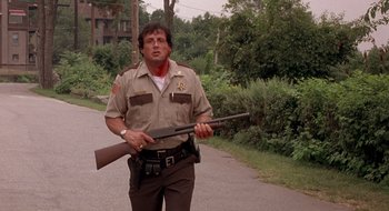 Movie still from “Cop Land” (1997), directed by James Mangold – A man holding a rifle on the side of a road; Medium shot, Low angle