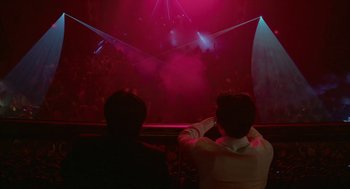 Movie still from “Cosmopolis” (2012), directed by David Cronenberg – Two people sitting in front of a stage with a projector; Wide shot, Low angle