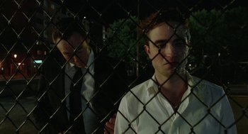 Movie still from “Cosmopolis” (2012), directed by David Cronenberg – A man standing behind a chain link fence with another man standing behind him; Medium shot, Over the shoulder angle