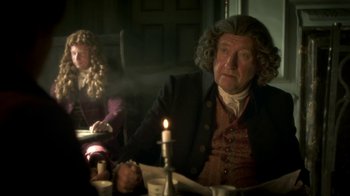 Movie still from “Crooked House” (2017), directed by Gilles Paquet-Brenner – A man sitting at a table with a candle in the background; Medium shot, Over the shoulder angle
