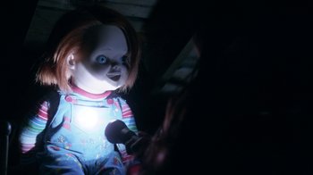 Movie still from “Curse of Chucky” (2013), directed by Don Mancini – A creepy doll with blue eyes is holding a microphone in a dark room; Medium shot, Overhead angle