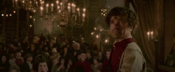 Movie still from “Cyrano” (2021), directed by Joe Wright – A crowd of people sitting in a room with lights; Medium shot, Over the shoulder angle