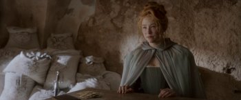 Movie still from “Cyrano” (2021), directed by Joe Wright – A woman sitting at a table in front of a stone wall; Medium shot, Over the shoulder angle