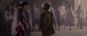 Movie still from “Cyrano” (2021), directed by Joe Wright – A group of men in medieval garb standing in a room; Medium shot, Over the shoulder angle