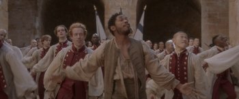 Movie still from “Cyrano” (2021), directed by Joe Wright – A group of men standing next to each other in front of a flag; Medium shot, Over the shoulder angle