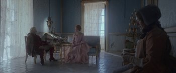 Movie still from “Cyrano” (2021), directed by Joe Wright – A woman in a pink dress sitting on a couch; Wide shot, Over the shoulder angle