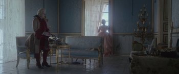 Movie still from “Cyrano” (2021), directed by Joe Wright – A woman standing in front of a window looking outside; Wide shot, High angle