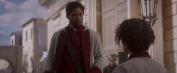 Movie still from “Cyrano” (2021), directed by Joe Wright – A man in a red and white jacket standing in front of a building; Medium shot, Over the shoulder angle