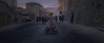Movie still from “Cyrano” (2021), directed by Joe Wright – A woman dressed in a long dress is standing in the middle of the street; Wide shot, Over the shoulder angle