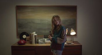 Movie still from “Flower” (2017), directed by Max Winkler – A woman standing in front of a desk holding a book; Medium shot, Over the shoulder angle