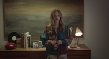 Movie still from “Flower” (2017), directed by Max Winkler – A woman standing in front of a desk holding a bottle; Medium shot, High angle