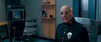 Movie still from “Demolition Man” (1993), directed by Marco Brambilla – A man in a green uniform and glasses in a room; Close Up shot, Over the shoulder angle