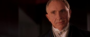 Movie still from “Demolition Man” (1993), directed by Marco Brambilla – An older man wearing a suit and tie; Close Up shot, Over the shoulder angle