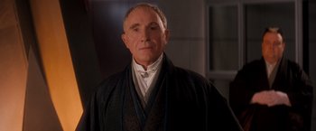 Movie still from “Demolition Man” (1993), directed by Marco Brambilla – An older man wearing a black robe and a white shirt; Close Up shot, Over the shoulder angle