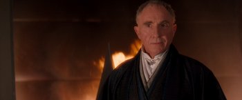 Movie still from “Demolition Man” (1993), directed by Marco Brambilla – An older man wearing a suit standing in front of a fire; Close Up shot, Over the shoulder angle