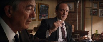 Movie still from “Ford v Ferrari” (2019), directed by James Mangold – A man wearing a suit and tie sitting at a table; Medium shot, Over the shoulder angle