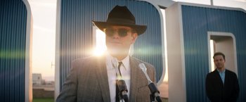 Movie still from “Ford v Ferrari” (2019), directed by James Mangold – A man in a suit and a hat standing in front of microphones; Medium shot, Low angle