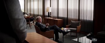 Movie still from “Ford v Ferrari” (2019), directed by James Mangold – An older man in a suit sitting in a chair in front of a table; Wide shot, Over the shoulder angle