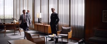 Movie still from “Ford v Ferrari” (2019), directed by James Mangold – A man in a suit standing next to a glass table; Wide shot, Low angle