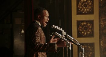 Movie still from “Detroit” (2017), directed by Kathryn Bigelow – A man in a brown jacket is speaking into microphones; Medium shot, Low angle