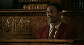 Movie still from “Detroit” (2017), directed by Kathryn Bigelow – A man wearing a suit and tie sitting in a wooden bench; Medium shot, High angle