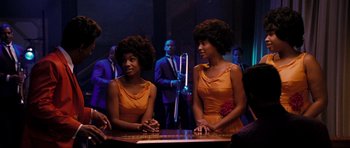 Movie still from “Dreamgirls” (2006), directed by Bill Condon – A couple of women sitting at a table in front of a microphone; Medium shot, Over the shoulder angle