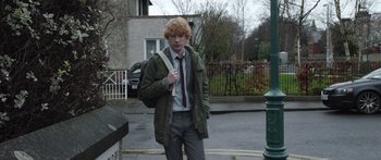 Movie still from “Frank” (2014), directed by Lenny Abrahamson – A man with a red hair and a green jacket is standing on the sidewalk; Medium shot, Over the shoulder angle