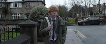 Movie still from “Frank” (2014), directed by Lenny Abrahamson – A young man in a tie and a jacket walking down a sidewalk; Medium shot, Over the shoulder angle