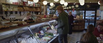 Movie still from “Frank” (2014), directed by Lenny Abrahamson – A man in a green jacket standing in front of a display case of food; Wide shot, Over the shoulder angle