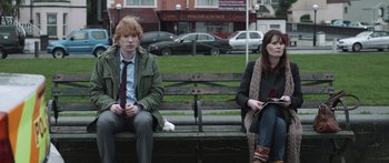 Movie still from “Frank” (2014), directed by Lenny Abrahamson – A man and a woman sitting on top of a wooden bench; Medium shot, Over the shoulder angle