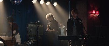 Movie still from “Frank” (2014), directed by Lenny Abrahamson – A group of people standing on a stage with lights in the background; Medium shot, Over the shoulder angle