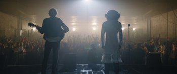 Movie still from “Frank” (2014), directed by Lenny Abrahamson – Two people standing in front of a crowd of people; Medium shot, Low angle
