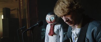 Movie still from “Frank” (2014), directed by Lenny Abrahamson – A person with a dummy head standing in front of a microphone; Close Up shot, Over the shoulder angle